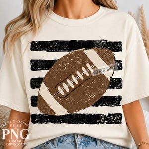 Op de afbeelding: Een crèmekleurig T-shirt met korte mouwen en een grafische print van een bruine American football, gecentreerd over horizontale zwarte, verweerde strepen. Het ontwerp bevat witte veters en een witte streep aan elk uiteinde met een textuur.