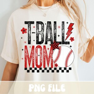 May include: Cream-colored t-shirt with the text "T-BALL MOM" in black and red. The design includes a baseball, a red lightning bolt, and floral accents. The shirt has a relaxed fit.