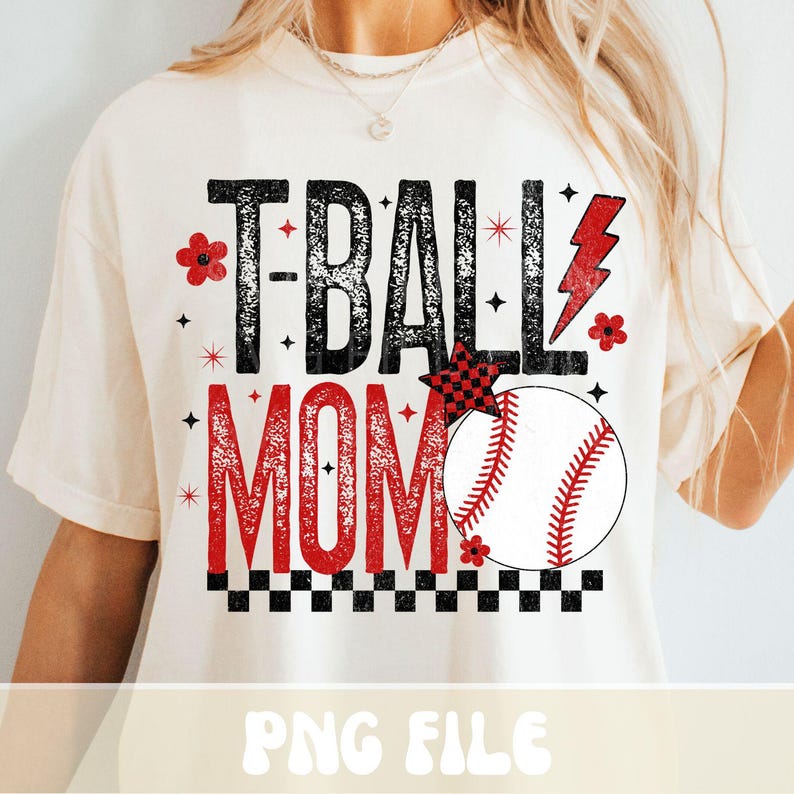May include: Cream-colored t-shirt with the text "T-BALL MOM" in black and red. The design includes a baseball, a red lightning bolt, and floral accents. The shirt has a relaxed fit.