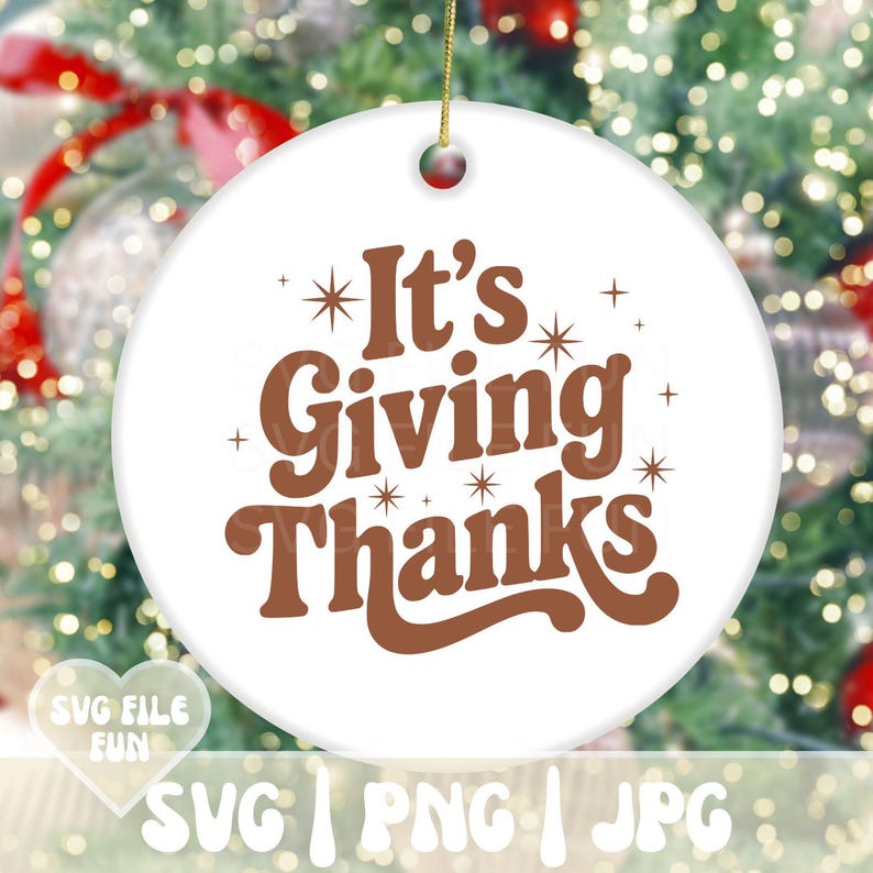 May include: A round white ceramic ornament with the text "It's Giving Thanks" in brown. The ornament has a gold string for hanging and is decorated with starbursts. The background is a blurred image of a Christmas tree.