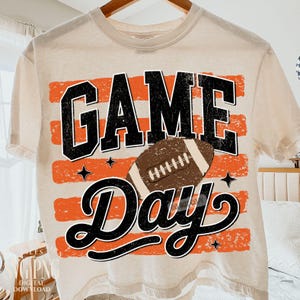 May include: A cream-colored t-shirt featuring a graphic print with the text GAME DAY in black, distressed block and script lettering. The design includes horizontal orange distressed stripes, a brown football, and small black star accents.