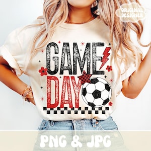 May include: Cream-colored t-shirt with the words "GAME DAY" in black and red, featuring a soccer ball graphic. The design includes a red lightning bolt and small floral accents. The shirt is a casual style.