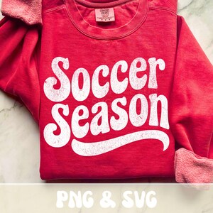Op de afbeelding: Een rode sweatshirt met de tekst "Soccer Season" in grote, witte letters in retrostijl. De sweatshirt heeft een ronde hals en lange mouwen met geribde manchetten. De afbeelding bevat ook de tekst "PNG & SVG".