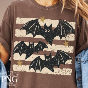 May include: A brown short-sleeve t-shirt featuring a graphic print of three black bats with textured wings. The design includes horizontal distressed cream-colored stripes and small gold glitter stars scattered across the front of the garment.