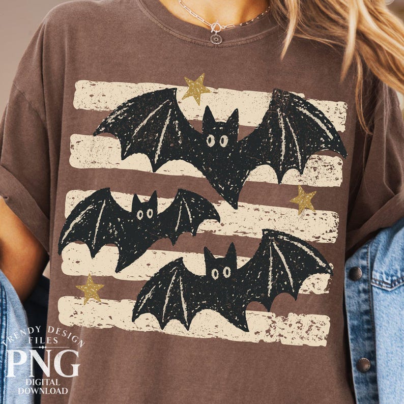 May include: A brown short-sleeve t-shirt featuring a graphic print of three black bats with textured wings. The design includes horizontal distressed cream-colored stripes and small gold glitter stars scattered across the front of the garment.