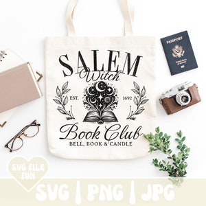 K&ouml;nnte beinhalten: Eine wei&szlig;e Einkaufstasche mit einem schwarzen Design, auf dem "Salem Witch Book Club Est. 1692 Bell, Book & Candle" steht. Das Design zeigt ein Buch, aus dem ein Mond und Sterne herauskommen.
