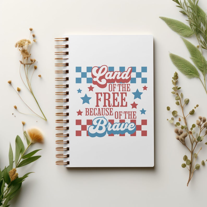 May include: White spiral-bound notebook with gold-colored coils. The cover features the text "Land of the Free Because of the Brave" in red, white, and blue, with star and checkerboard design elements. Decorative sprigs of dried flowers and greenery surround the notebook.