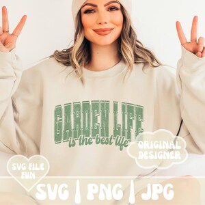 May include: A light beige sweatshirt with the text "GARDEN LIFE is the best life" in green lettering. The text is in a vintage style font. There is a white cloud with the text "ORIGINAL DESIGNER" in black lettering. The sweatshirt is being worn by a person with long blonde hair and a white beanie.