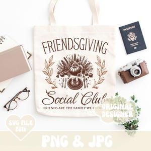 May include: A cream-colored tote bag with the words "FRIENDSGIVING Social Club" in brown script. The bag features a fall-themed graphic with a pumpkin, leaves, and cutlery. A passport, camera, and glasses are also visible.