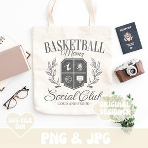 May include: A white tote bag with a black and gray design that says "Basketball Moms Social Club Loud and Proud". The design features a basketball, a basketball hoop, and a silhouette of a person playing basketball. The bag is on a white surface with a passport, a camera, and a plant.