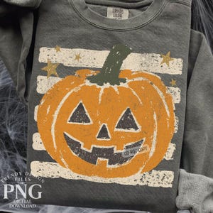 Puede incluir: Una sudadera de cuello redondo gris carbón con un gráfico de calabaza naranja desgastado centrado sobre rayas horizontales blancas y estrellas doradas. La prenda tiene una etiqueta de la marca Comfort Colors en el cuello interior.