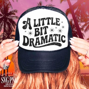 May include: Black and white trucker hat with the text "A Little Bit Dramatic" in a bold, retro font. The hat has a mesh back and a curved brim. The design includes star accents.