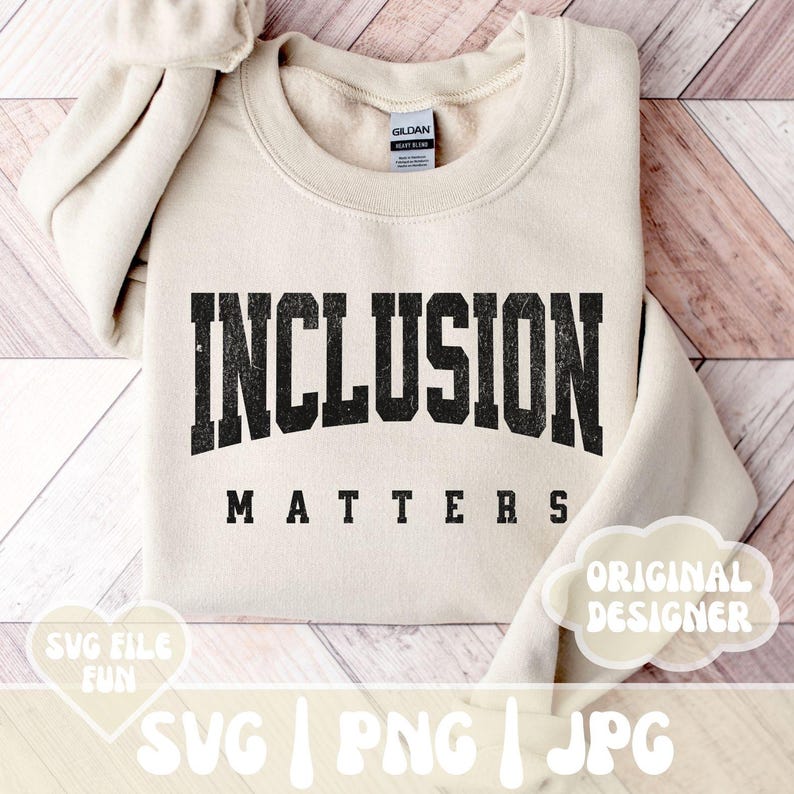 May include: A beige sweatshirt with the text "INCLUSION MATTERS" in black with a distressed look. The text is in a large, bold font. The sweatshirt is on a wooden surface.