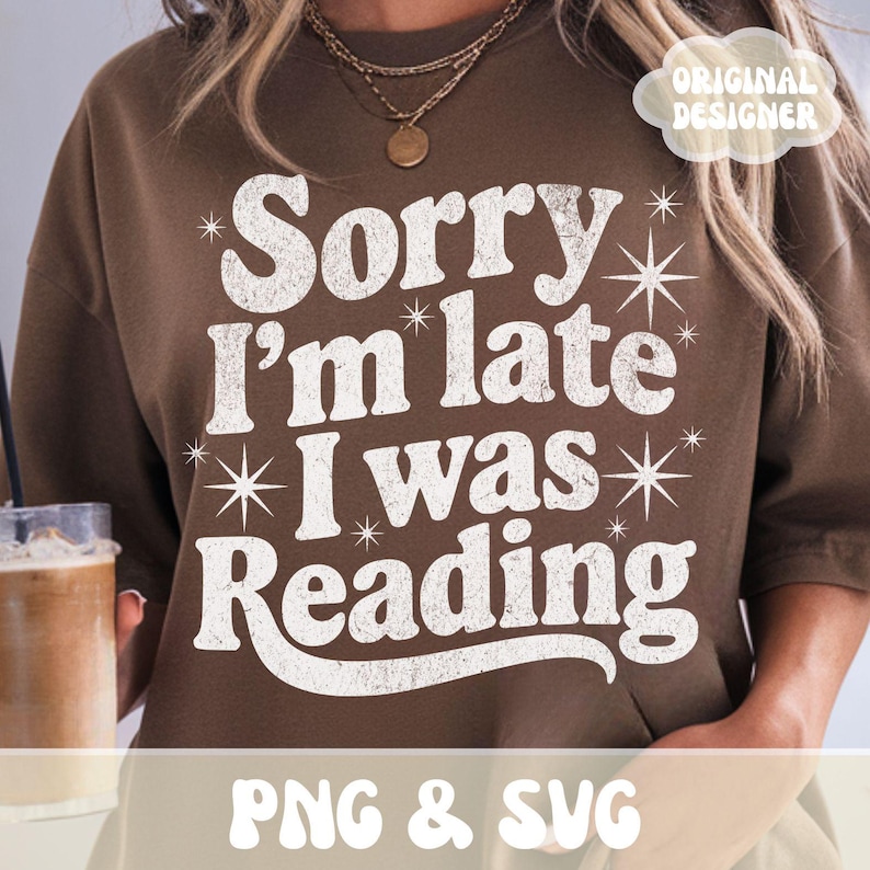 Op de afbeelding: Een bruine sweatshirt met de witte tekst "Sorry I'm late I was Reading", versierd met sterren. Het ontwerp heeft een verweerde, vintage uitstraling. De woorden "PNG & SVG" staan onderaan.