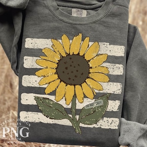 May include: A charcoal gray crewneck sweatshirt featuring a large, yellow sunflower graphic with a dark brown center and green leaves. Behind the sunflower is a horizontal, distressed white striped pattern. The inner neck label reads Comfort Colors.