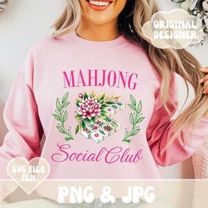 Puede incluir: Sudadera rosa con el texto "Mahjong Social Club" en letras rosas. Un dise&ntilde;o floral con fichas de mahjong blancas est&aacute; por encima del texto.