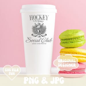May include: White coffee cup with a black and white graphic design that says "Hockey Moms Social Club Loud and Proud". The graphic features a hockey helmet, crossed hockey sticks, and a glass of wine. The cup is sitting on a pink surface with a stack of colorful macarons.