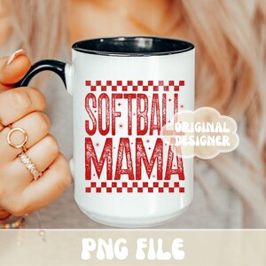 May include: White ceramic mug with a black handle and rim. The mug features the words "SOFTBALL MAMA" in red, with a checkered pattern above and below the text. The mug is held by a person.