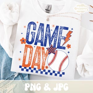 Pode incluir: Camisola branca com as palavras "GAME DAY" em azul e laranja, juntamente com um gráfico de basebol. O design inclui uma bola de basebol, um raio e detalhes de estrelas. As palavras "PNG & JPG" estão na parte inferior.