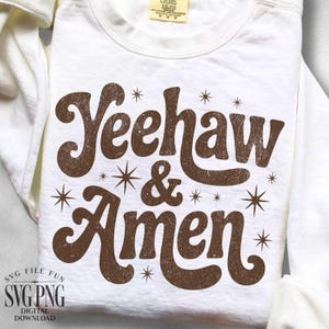 Puede incluir: Sudadera blanca con el texto "Yeehaw & Amen" en marr&oacute;n, con letras de estilo retro. El dise&ntilde;o incluye detalles de estrellas. La sudadera est&aacute; doblada, con las mangas visibles.