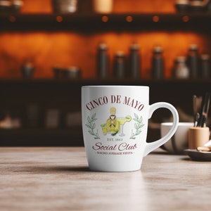 Puede incluir: Taza de cer&aacute;mica blanca con un dise&ntilde;o de Cinco de Mayo. El dise&ntilde;o incluye texto e ilustraciones de una margarita, una botella de tequila y las palabras "Social Club". La taza tiene un asa curva y est&aacute; sobre una superficie de madera.