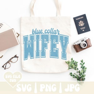 Op de afbeelding: Een cr&egrave;mekleurige canvas draagtas met de tekst blue collar WIFEY in een verweerd lichtblauw collegiaal lettertype. De tas is omringd door een notitieboek, een bril, een paspoort, een vintage camera en een kleine groene tak met blaadjes.