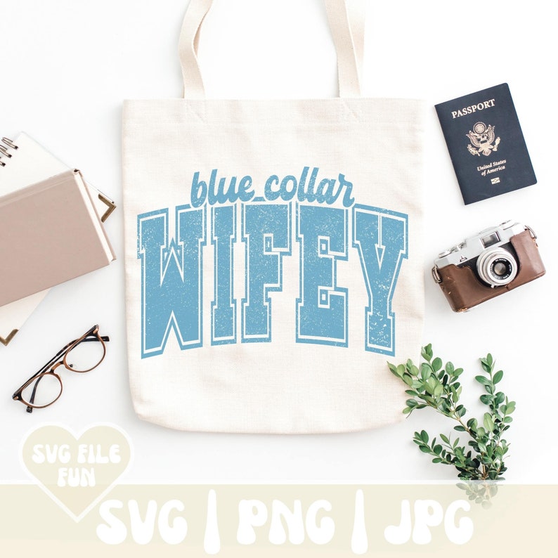 Op de afbeelding: Een cr&egrave;mekleurige canvas draagtas met de tekst blue collar WIFEY in een verweerd lichtblauw collegiaal lettertype. De tas is omringd door een notitieboek, een bril, een paspoort, een vintage camera en een kleine groene tak met blaadjes.