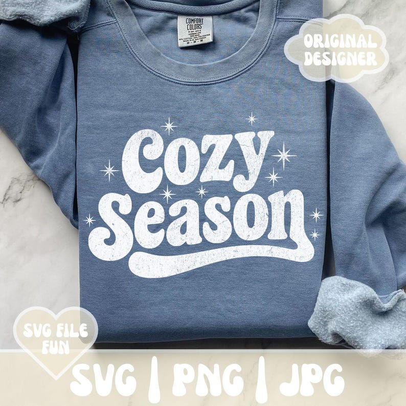 Puede incluir: Sudadera azul Comfort Colors con las palabras "Cozy Season" en blanco, con letras de estilo retro. El dise&ntilde;o incluye detalles de estrellas. Tambi&eacute;n se ven las palabras "Original Designer" y "SVG File Fun".