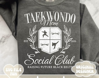 May include: A gray sweatshirt with white text that reads "Taekwondo Mom Social Club Raising Future Black Belts" with a crest featuring a taekwondo pose and the years "EST. 2025".