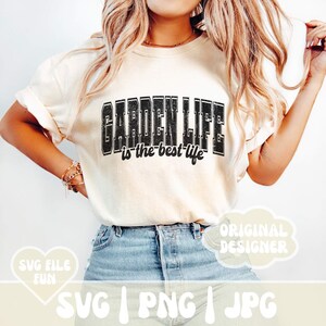 May include: A white t-shirt with the text "GARDEN LIFE is the best life" in black lettering. The text has a distressed, vintage look. The t-shirt is being worn by a person with long blonde hair and blue jeans.