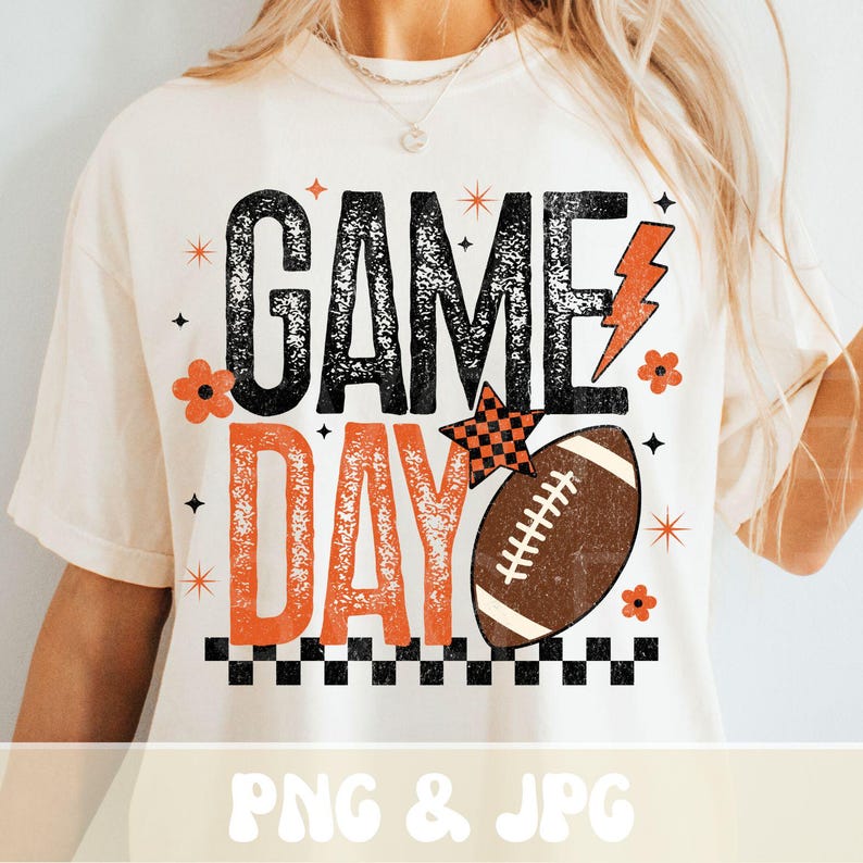 May include: Cream-colored t-shirt with a retro "Game Day" design. The design features the words "Game Day" in black and orange, a football, and decorative elements like stars and flowers. The shirt has a relaxed fit.