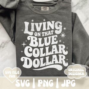 May include: A dark gray sweatshirt with the text "Living on that Blue Collar Dollar" in white, retro-style lettering. The design includes starburst and star accents. The sweatshirt also has the words "SVG File Fun" and "Original Designer" on the bottom.