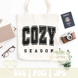 May include: A white tote bag with black text that reads "Cozy Season". The text is in a distressed font and has a vintage look. The bag is sitting on a white surface.