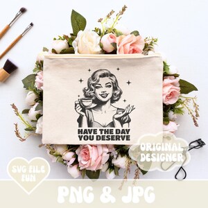 Puede incluir: Un neceser de lona beige con una ilustraci&oacute;n en blanco y negro de una mujer sosteniendo una taza de caf&eacute;, con el texto "HAVE THE DAY YOU DESERVE". La bolsa est&aacute; rodeada de rosas rosas y blancas y vegetaci&oacute;n, con brochas de maquillaje en el fondo.