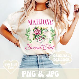 Puede incluir: Una camiseta blanca con el texto "Mahjong Social Club" en letras rosas. Un dise&ntilde;o floral con flores rosas y hojas verdes rodea una pila de fichas de mahjong.