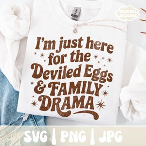 Puede incluir: Sudadera blanca con el texto "I'm just here for the Deviled Eggs & FAMILY DRAMA" en letras marrones. El dise&ntilde;o incluye acentos de estrellas. La sudadera es de la marca Gildan.