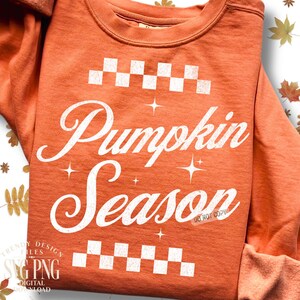 May include: Orange sweatshirt with the words "Pumpkin Season" in white script. The design includes white checkered patterns above and below the text, with star accents. The sweatshirt has long sleeves.