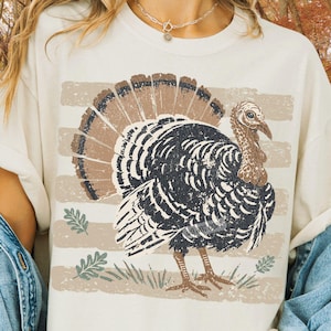 May include: A cream-colored t-shirt featuring a distressed graphic of a turkey with a fanned tail. The turkey is black and brown, positioned over horizontal beige stripes and small green leaf illustrations. The design has a weathered, vintage texture.