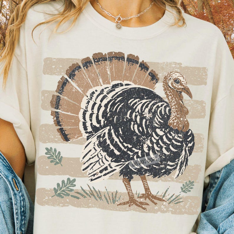 May include: A cream-colored t-shirt featuring a distressed graphic of a turkey with a fanned tail. The turkey is black and brown, positioned over horizontal beige stripes and small green leaf illustrations. The design has a weathered, vintage texture.