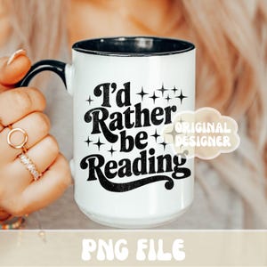 Op de afbeelding: Witte keramische mok met een zwarte binnenkant en handvat. De mok heeft de tekst "I'd Rather be Reading" in een retro lettertype. De mok wordt vastgehouden door een persoon die ringen draagt.