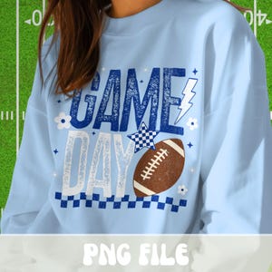Puede incluir: Sudadera azul claro con las palabras "GAME DAY" en azul y blanco, con un bal&oacute;n de f&uacute;tbol americano marr&oacute;n. El dise&ntilde;o incluye una estrella a cuadros, un rayo y flores. La parte inferior tiene un borde a cuadros.