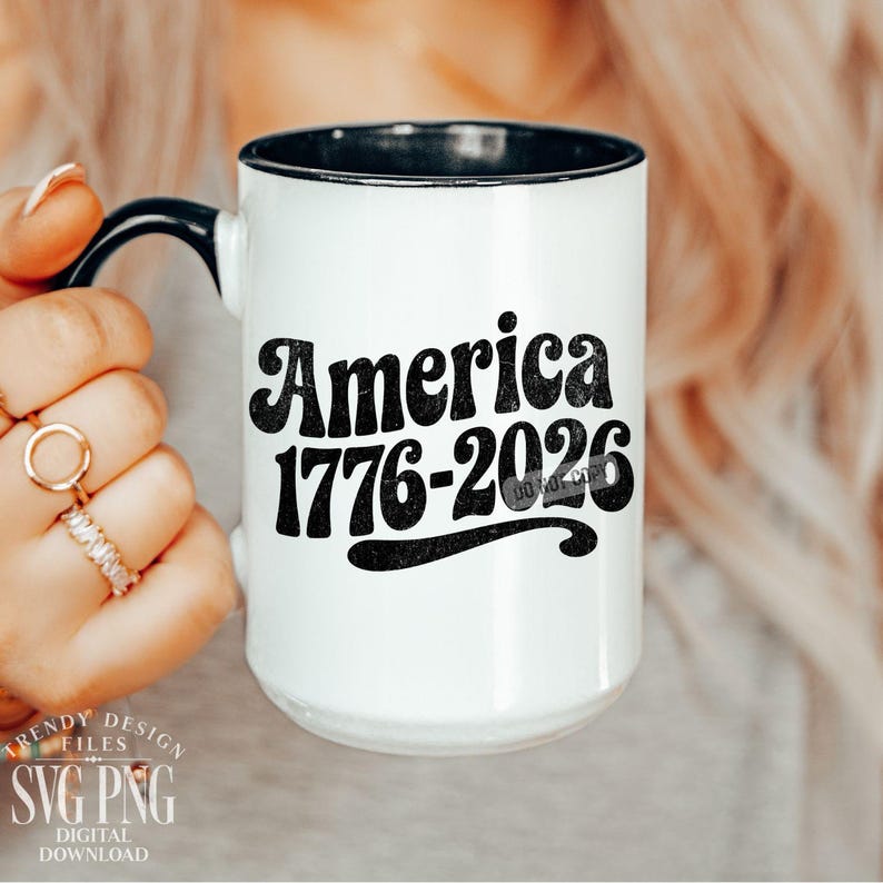 May include: White ceramic mug with a black rim and handle. The mug features the text "America 1776-2026" in a bold, retro font. The mug is held by a person with rings on their fingers. The mug is a patriotic item.