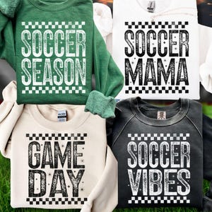 K&ouml;nnte beinhalten: Vier Sweatshirts mit Fu&szlig;ball-Text. Die Sweatshirts sind in Gr&uuml;n, Wei&szlig;, Creme und Anthrazitgrau gehalten. Die Texte lauten "SOCCER SEASON", "SOCCER MAMA", "GAME DAY" und "SOCCER VIBES". Jedes Design hat einen karierten Rand.
