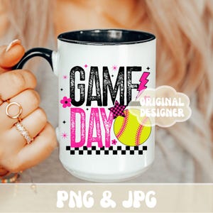 May include: White ceramic mug with a black handle and rim, featuring the words "GAME DAY" in black and pink, a yellow softball, and a pink lightning bolt. The mug has a black and white checkered pattern at the bottom.