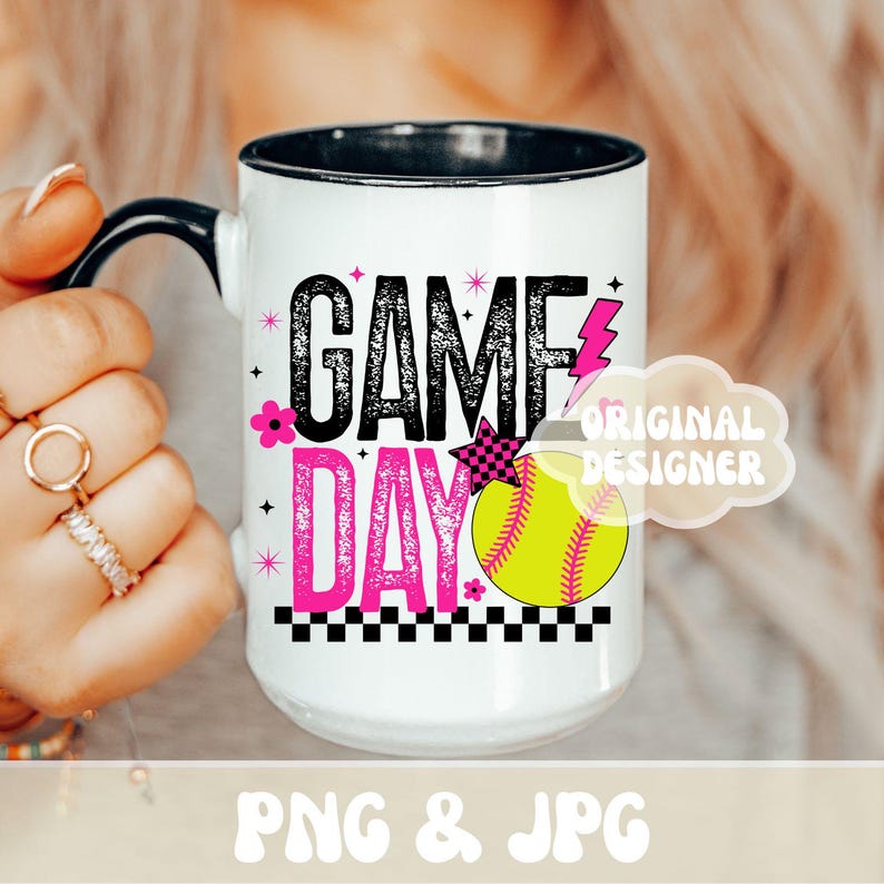 May include: White ceramic mug with a black handle and rim, featuring the words "GAME DAY" in black and pink, a yellow softball, and a pink lightning bolt. The mug has a black and white checkered pattern at the bottom.