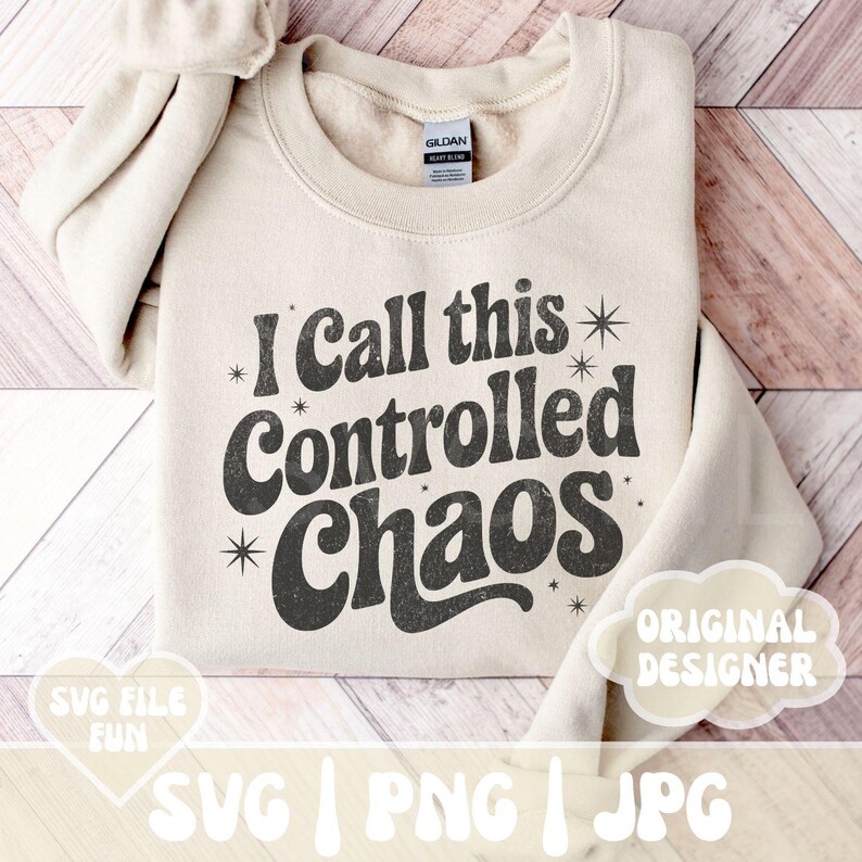 May include: A beige sweatshirt with the text "I Call this Controlled Chaos" in a retro font. The design includes starburst accents. The sweatshirt is folded on a wooden surface.