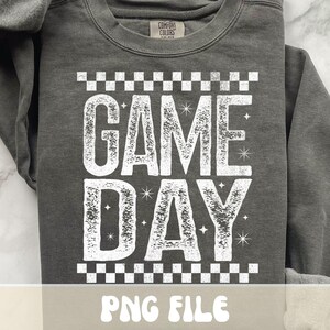 K&ouml;nnte beinhalten: Dunkelgraues Sweatshirt mit dem Schriftzug "GAME DAY" in gro&szlig;en, verblichenen wei&szlig;en Buchstaben. Der Text wird von einem Schachbrettmuster oben und unten eingerahmt. Das Sweatshirt hat einen Rundhalsausschnitt und lange &Auml;rmel.