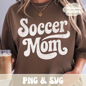 Op de afbeelding: Een bruine sweatshirt met de tekst "Soccer Mom" in een witte retro-lettertype. De sweatshirt heeft een ronde hals en korte mouwen. De tekst "PNG & SVG" is ook zichtbaar.