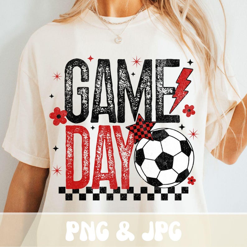 May include: Cream-colored t-shirt with a soccer-themed graphic. The design features the words "GAME DAY" in black and red, a soccer ball, a red lightning bolt, and decorative elements. The shirt is a casual wear item.