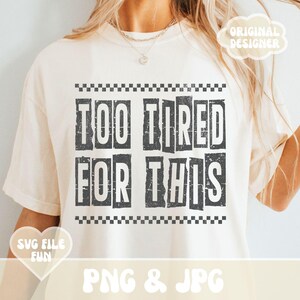 以下が含まれることがあります： 黒い文字で「Too Tired For This」と書いてある白いTシャツ。文字はかすれたようなフォントで、黒と白の市松模様の枠で囲まれている。